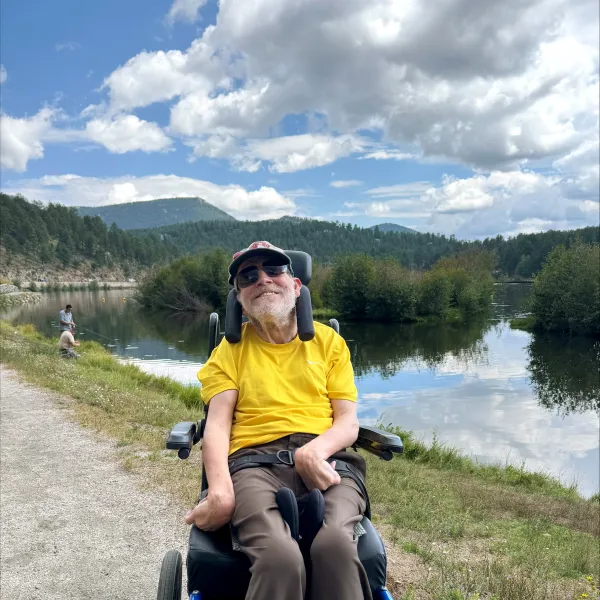 Smiling man in a yellow shirt sitting in a blue wheelchair by a river with trees and mountains under a partly cloudy sky.