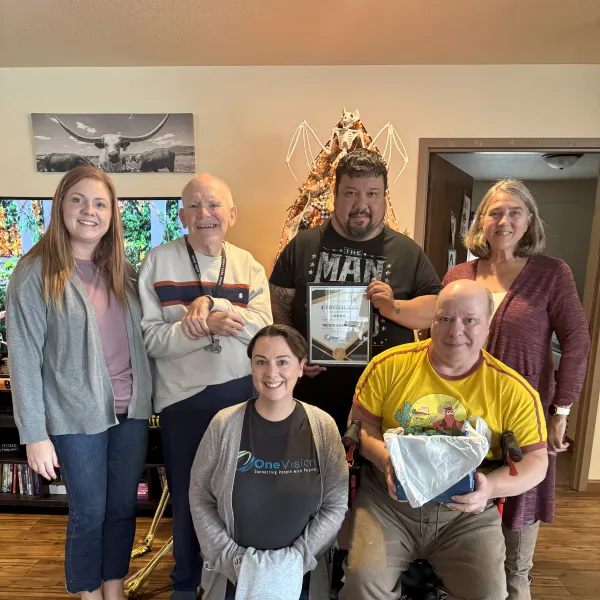 Group of six diverse adults smiling in a cozy living room with certificate and gifts on wooden floor.