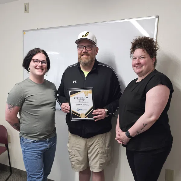 Three people smiling indoors, one holding a certificate, standing in front of a whiteboard and wall outlet.