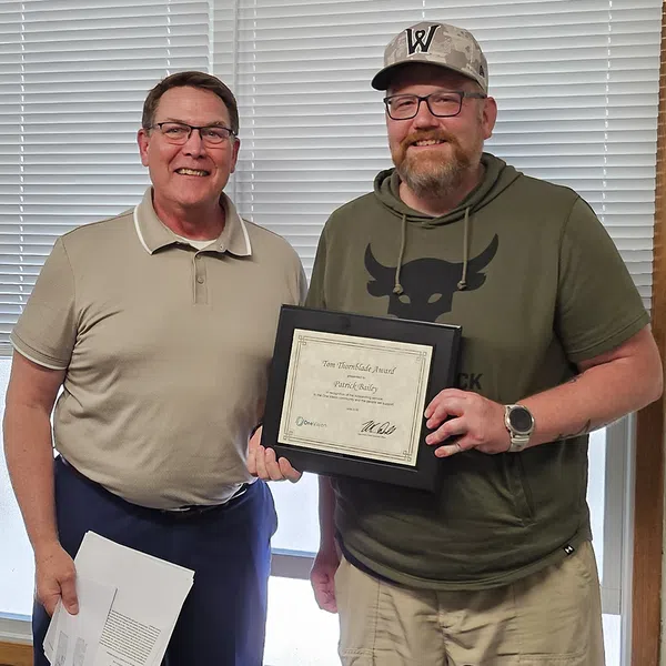 Two men smiling indoors, one holding a framed certificate of achievement in front of closed blinds.