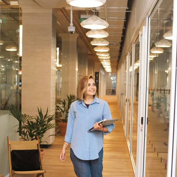 Smiling woman in blue shirt walking down bright modern office hallway holding tablet device.