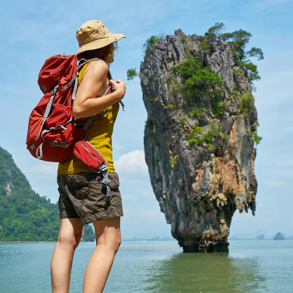 Hiker with red backpack and beige hat admiring tall limestone karst island in green water under blue sky.