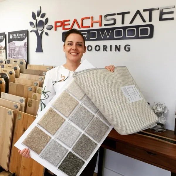 Smiling woman holding carpet and hardwood flooring samples inside a flooring showroom with Peach State Hardwood logo.
