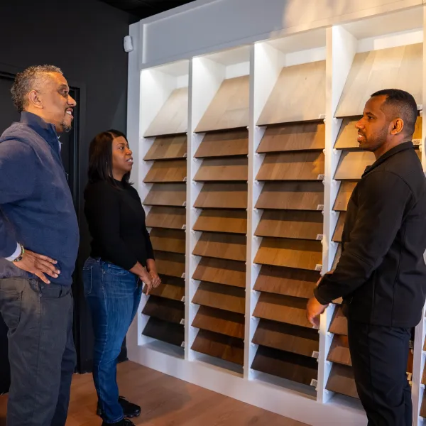 Three people discussing wood flooring samples displayed on a wall in a showroom setting.