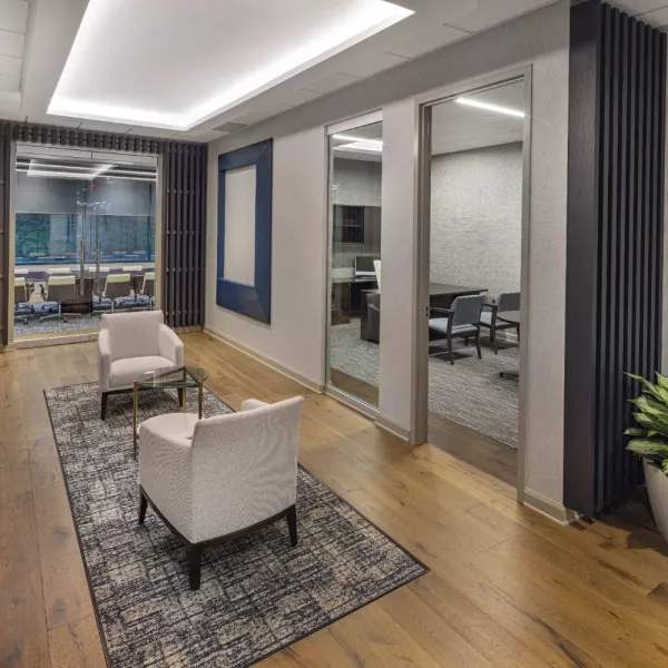 Modern office hallway with wooden floors, two white chairs on a patterned rug, and conference rooms with glass walls.