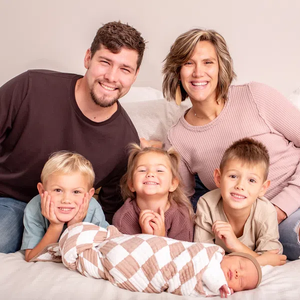Happy family of six including parents and four children posing together on a bed with a newborn baby.