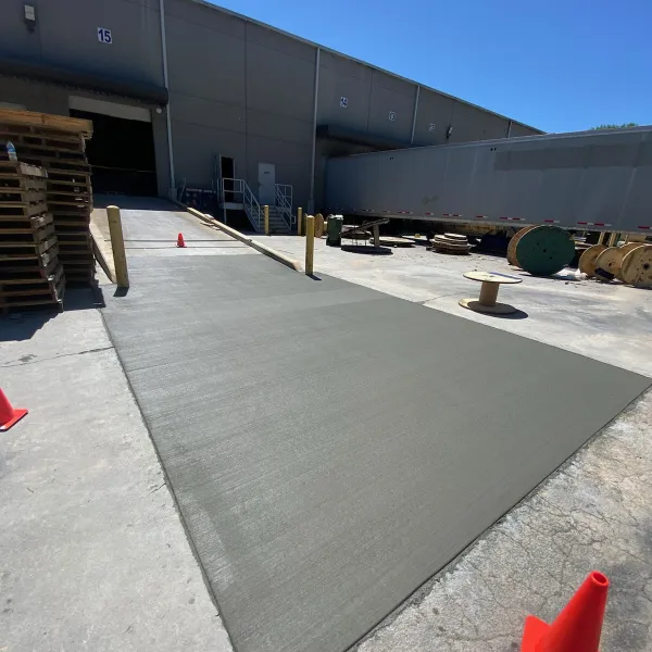 A loading dock with a new gray surface and safety cones, featuring pallets and spools in the background.