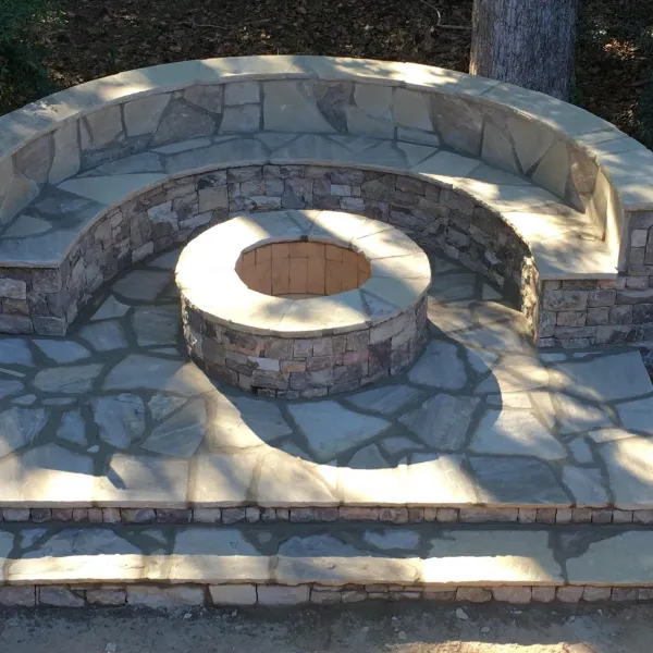 Stone fire pit with curved stone bench seating on a multi-level stone patio in a shaded outdoor area