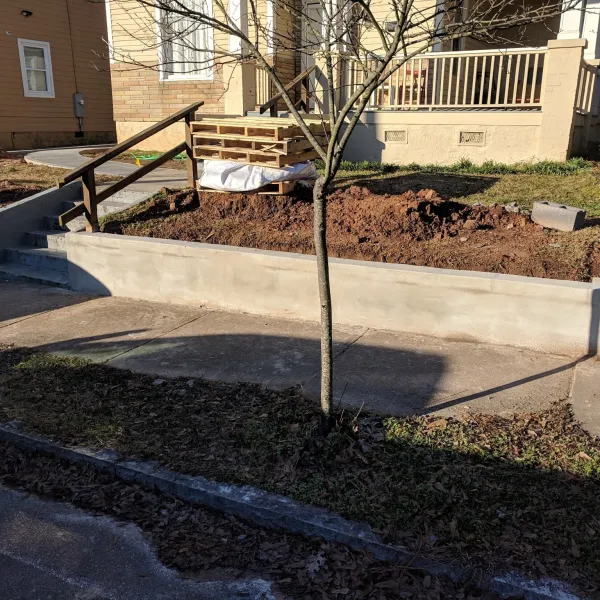 Newly constructed concrete retaining wall and stairs outside a residential house with bare soil and a small tree.