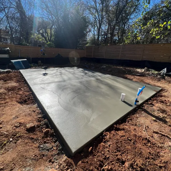 Freshly poured concrete slab foundation in a backyard with exposed soil and surrounding wooden fence under sunlight.