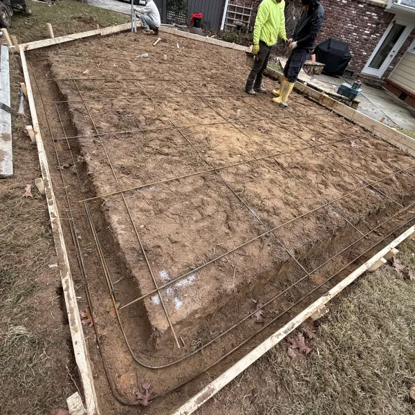 Home driveway under construction with steel rebar grid laid over gravel base next to a white house and shed.