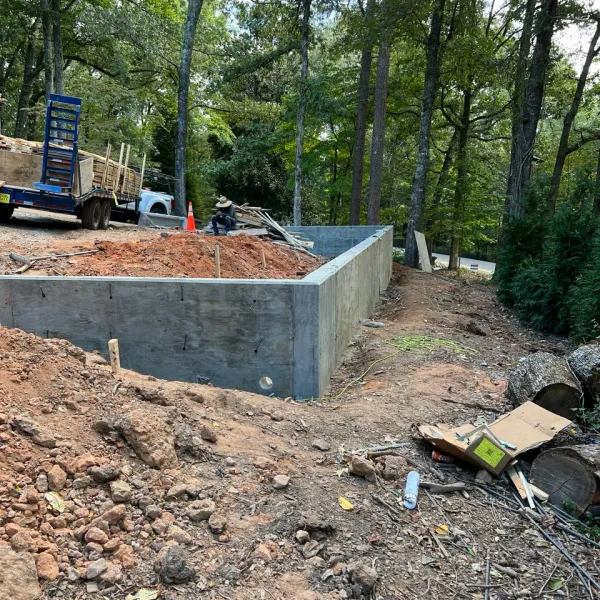 Construction site with concrete foundation walls surrounded by dirt, wood logs, and trees in a forested area.