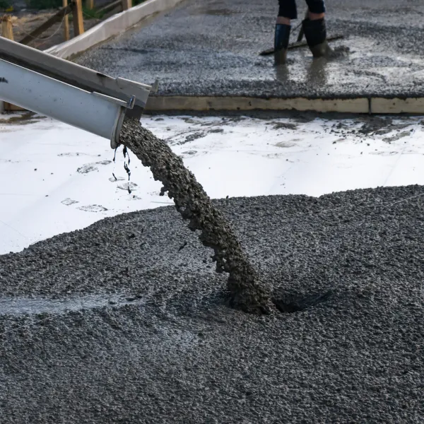 Wet concrete being poured from a chute onto a construction site floor with a worker in boots in the background.