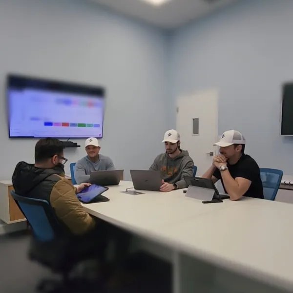 Four men in casual clothing and hats working on laptops and tablets in a modern conference room with wall screens.