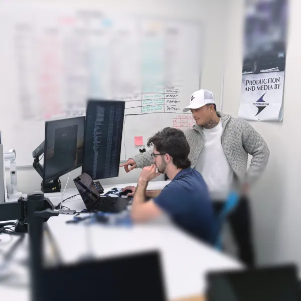 Two men collaborate on coding at a desk with multiple monitors and a whiteboard schedule in an office setting.