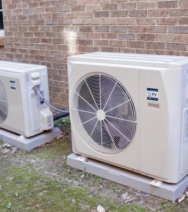 Two outdoor air conditioning units installed on concrete pads against a brick house wall with grassy ground.