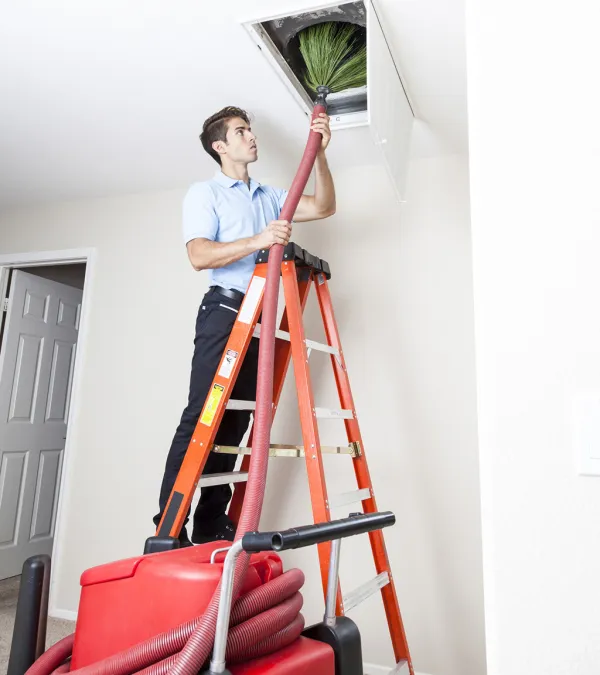 Technician on orange ladder cleaning ceiling air duct with vacuum and brush attachment in a residential room