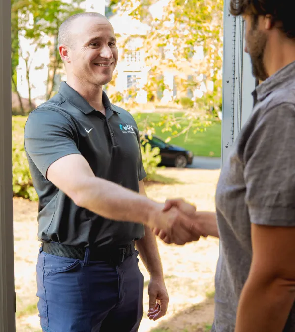 Two men shaking hands at a doorway, one in a black polo and the other in a gray shirt, outdoors in daylight
