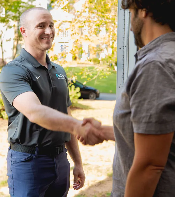 Two men shaking hands at a doorway, one in a black polo and the other in a gray shirt, outdoors in daylight