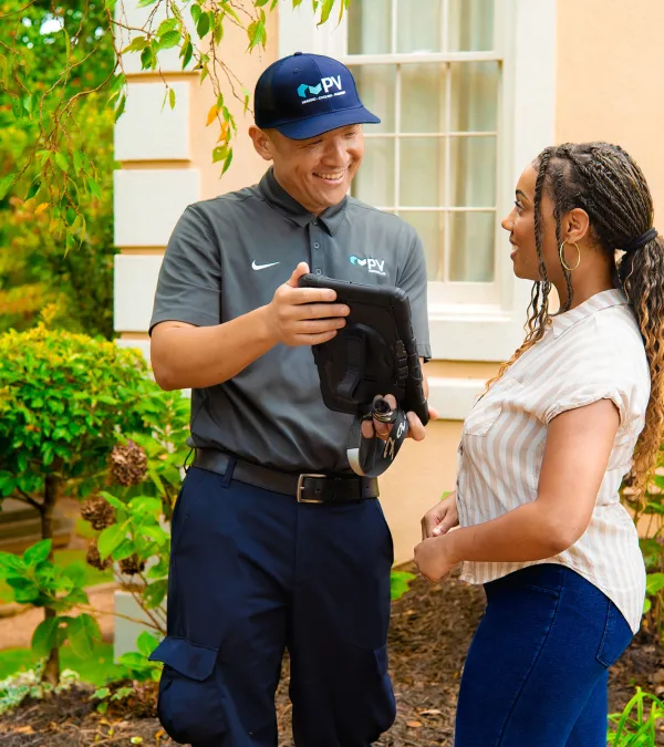 Technician in uniform showing a tablet to a woman outside a house with greenery and windows in the background