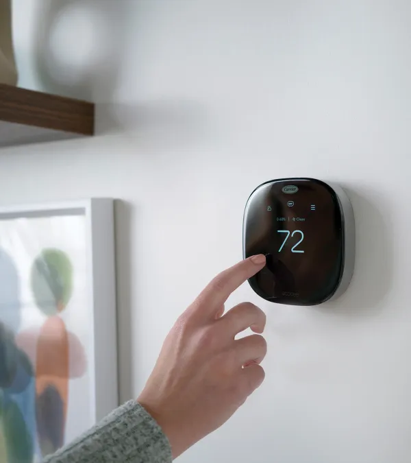 Person adjusting a modern black smart thermostat displaying 72 degrees mounted on a white wall.