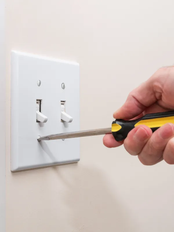 Hand using a yellow and black screwdriver to tighten a screw on a white double light switch plate on a beige wall.