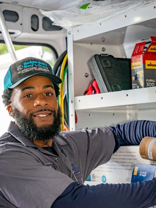 Smiling technician in uniform organizing tools and parts inside a service van with shelves and equipment.