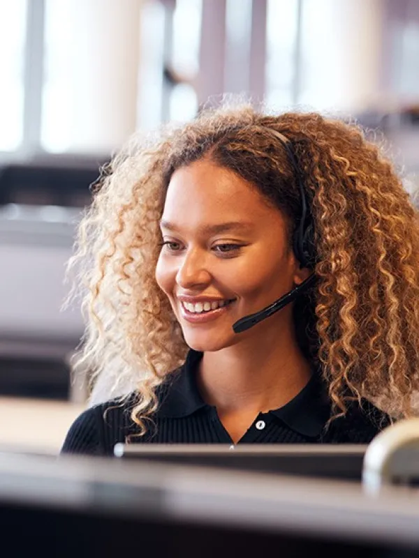 Man in blue shirt talking on phone while sitting at desk with computers and printer in office environment.
