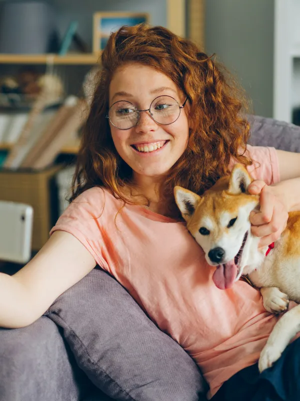 Smiling woman with glasses taking a selfie while holding a happy Shiba Inu dog on the couch at home
