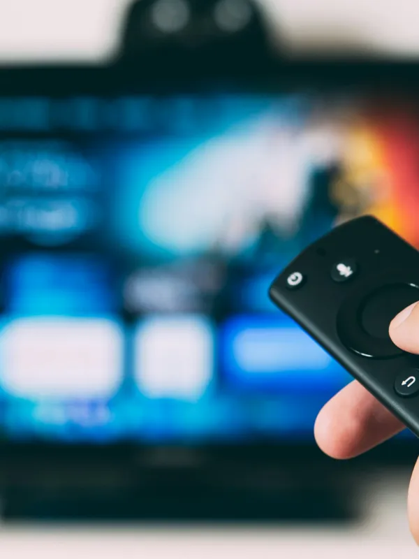 Hand holding a black remote control pointed at a blurred smart TV screen with streaming app icons.