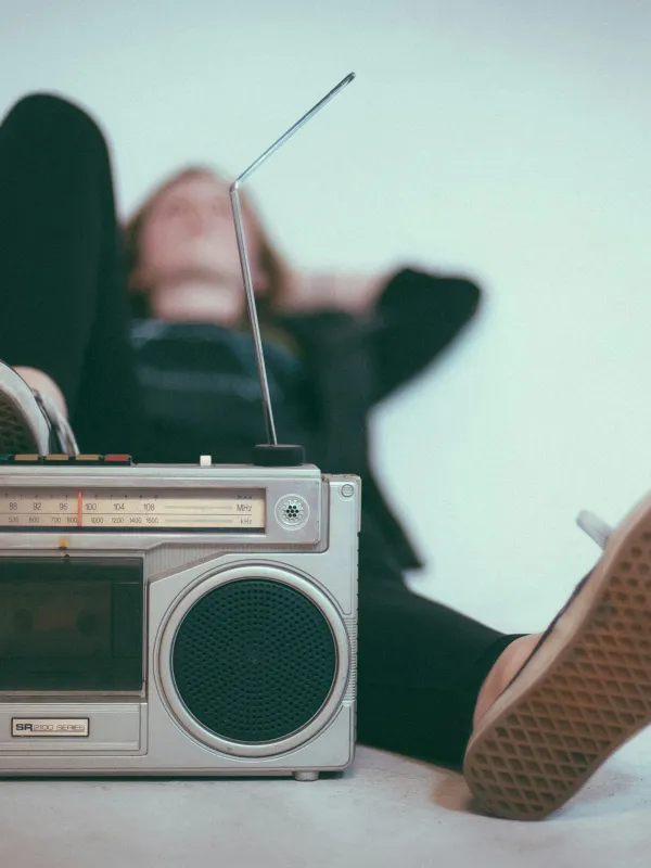 Vintage silver boombox with antenna in focus, person relaxing in background wearing black with sneakers visible.