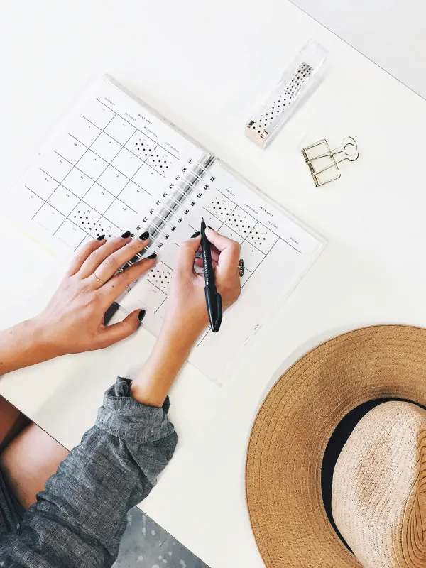 Person writing in a planner with a hat and stationery on a white desk.