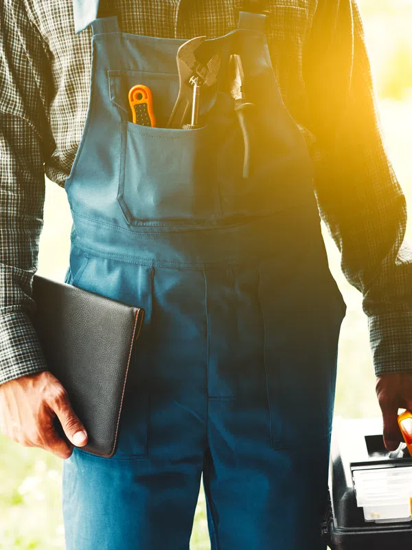 Man wearing blue overalls holding a toolbox and a tablet entering through a door with sunlight in the background
