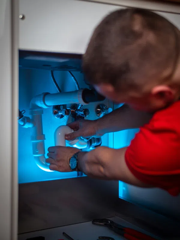 Plumber repairing white PVC pipes under a sink with blue lighting and tools nearby.