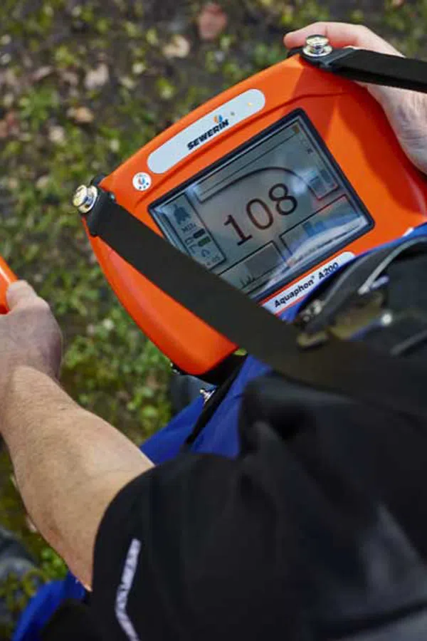 Person using orange underground pipe locator device with digital screen reading 108 in outdoor setting.