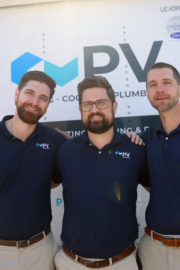 Three smiling men wearing matching PV company navy shirts standing in front of a service truck outdoors