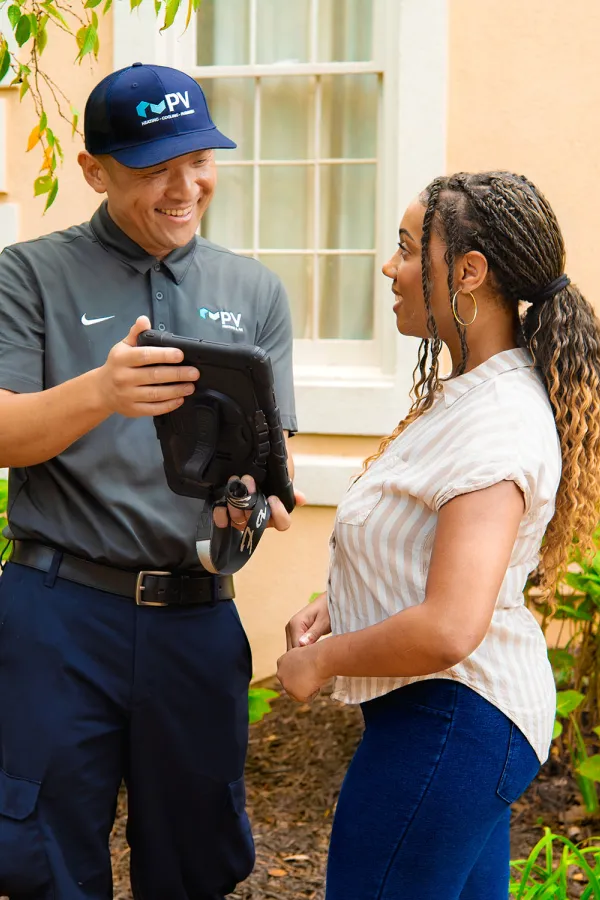 Technician in uniform showing a tablet to a woman outside a house with greenery and windows in the background