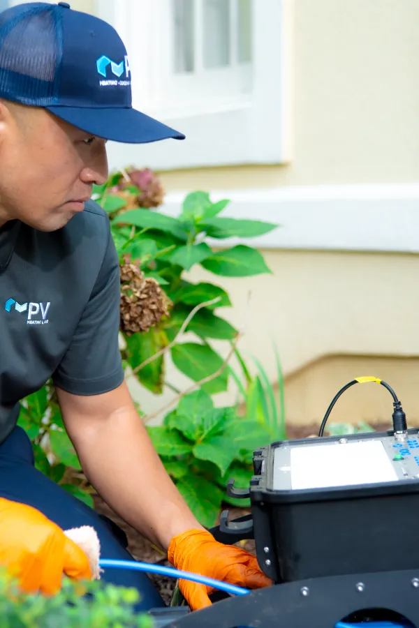 Technician in uniform and gloves using diagnostic equipment outdoors near a house wall and plants.