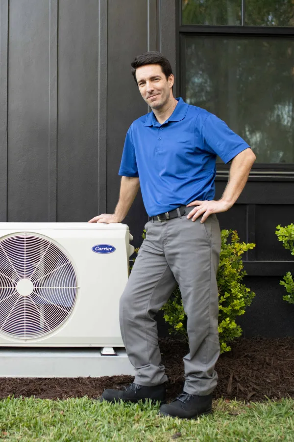 Technician standing next to a Carrier air conditioning unit outside a modern home with greenery.