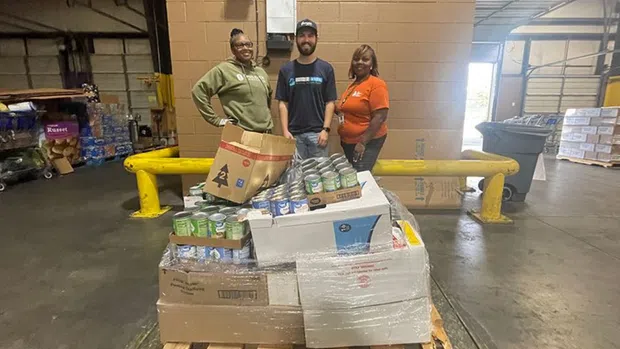 Three people standing behind a pallet loaded with canned goods and boxes inside a warehouse or distribution center.