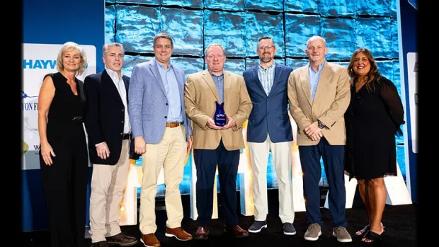 Seven professionals in formal attire posing on stage at an award event with a blue-lit background.
