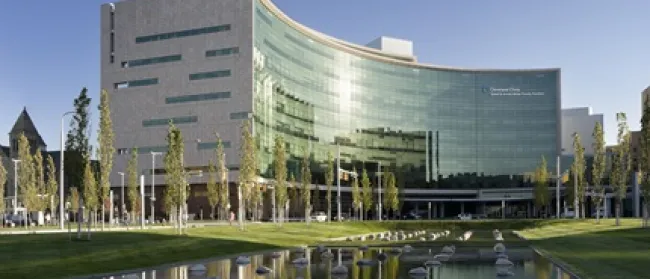 Modern curved glass hospital building with reflecting pond and trees in front during daytime.