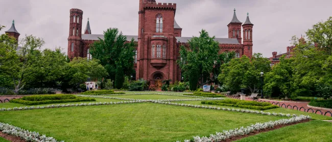 Red-brick historic castle building behind manicured gardens and green trees under cloudy sky