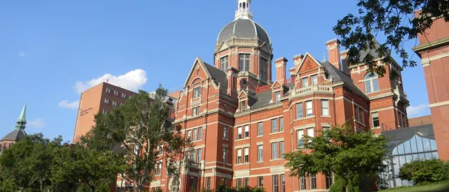 Historic red brick building with a large dome and spires under a clear blue sky, surrounded by trees and lawn.
