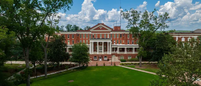 Historic red brick university building with white columns surrounded by green trees under cloudy blue sky.