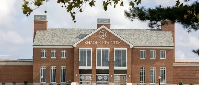 Front view of Shadek Stadium with brick facade, large windows, and stairs leading to the entrance under a partly cloudy sky.
