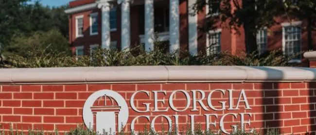 Georgia College brick sign with white pillars and red-brick campus building in the background under blue sky.