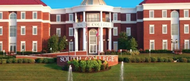 Red brick South College building with dome, surrounded by flags, fountains, and manicured lawn under clear sky