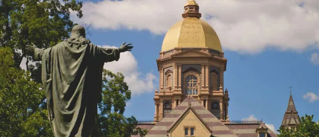 Statue of Jesus with outstretched arms facing golden dome of Notre Dame's Main Building under blue sky.