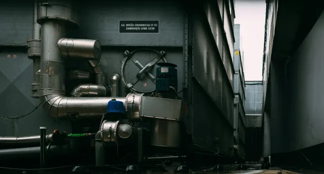 Industrial metal pipes and machinery with valves in a dimly lit narrow corridor or facility area.